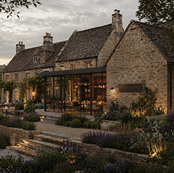 Stone cottage at dusk with warm interior lighting, stone steps, and a blooming garden in front.