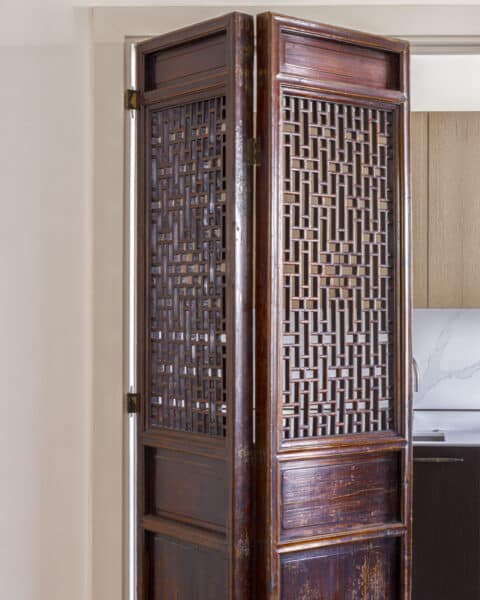 Pair of antique wooden folding doors with geometric lattice panels, opened to reveal a kitchen with light wood cabinetry behind.