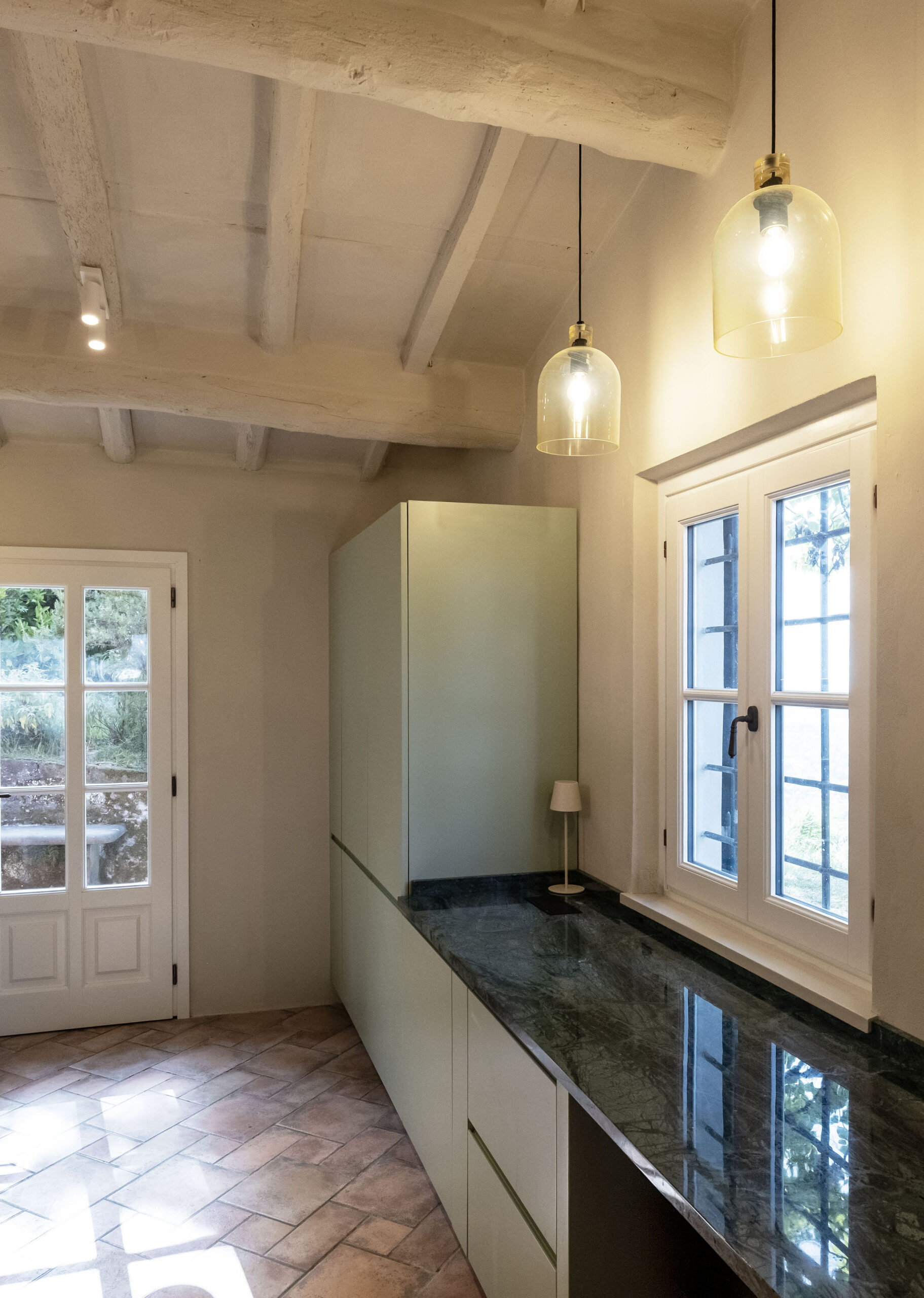 Kitchen interior with pale green cabinets along a dark marble countertop, white exposed ceiling beams, and glass pendant lights.