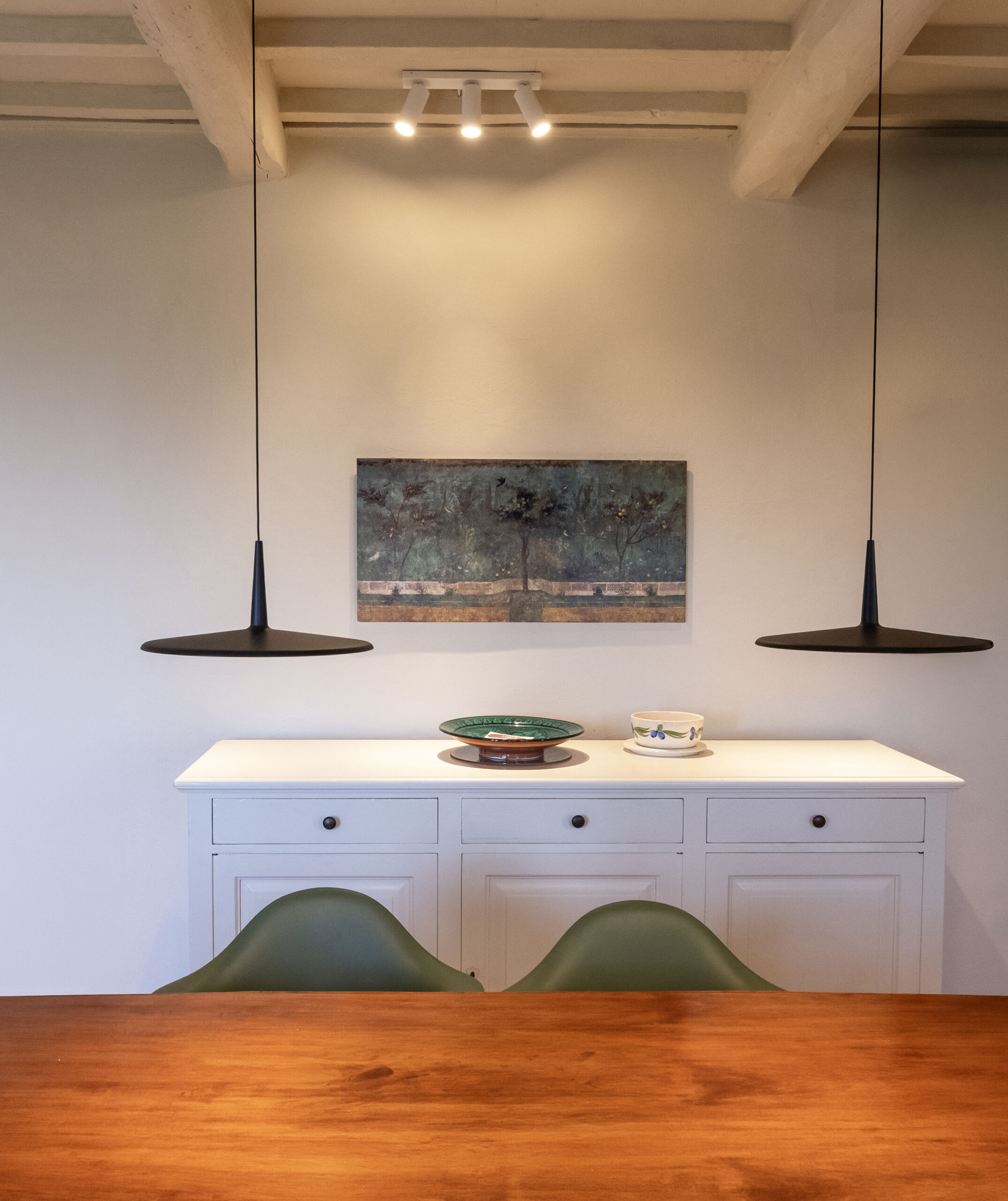 White cabinet with multiple drawers against a light wall, a landscape painting mounted above, and two black pendant lamps hanging from the ceiling, with a wooden table in the foreground and dishware on the white surface.