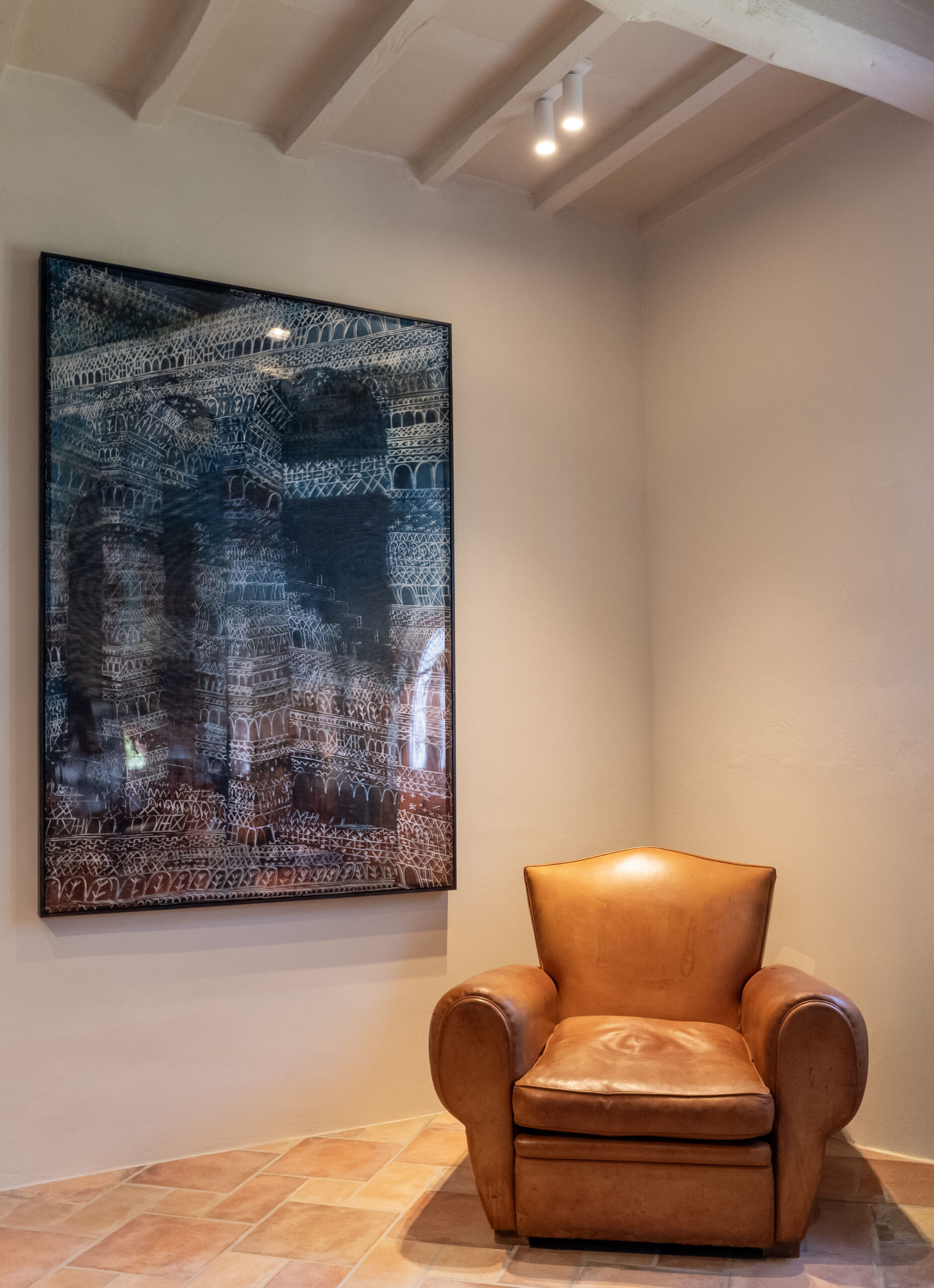Brown leather armchair in a corner beside a large framed blue architectural print on beige walls with ceiling beams above.