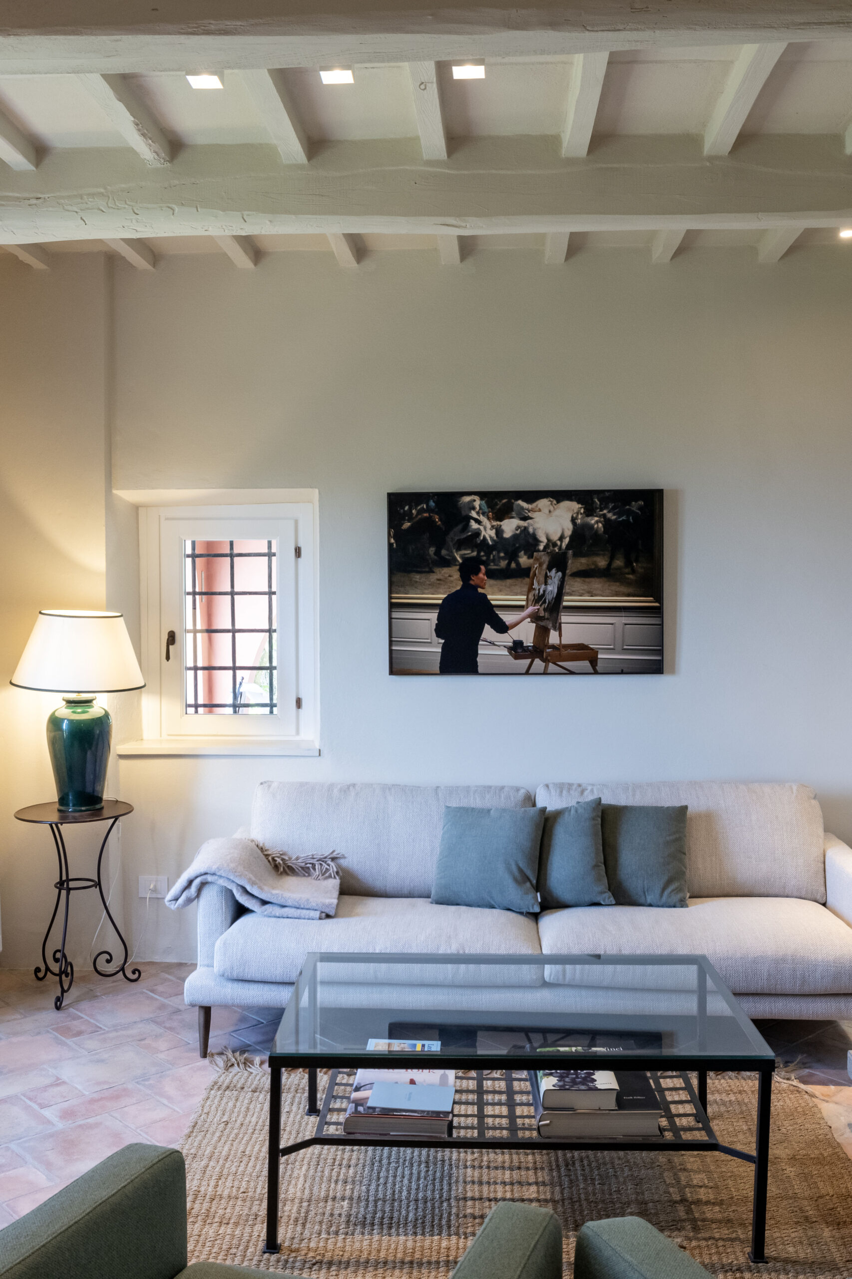 Cozy living room with a beige sofa, blue pillows, a glass coffee table, and a painting of a painter at an easel on the wall above.