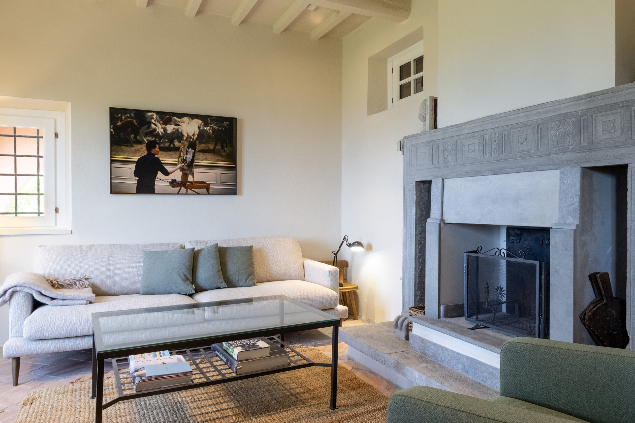 Living room with beige walls, light sofa and teal cushions, glass coffee table, painting of an artist at an easel, window left, and a large carved stone fireplace on the right attraction.