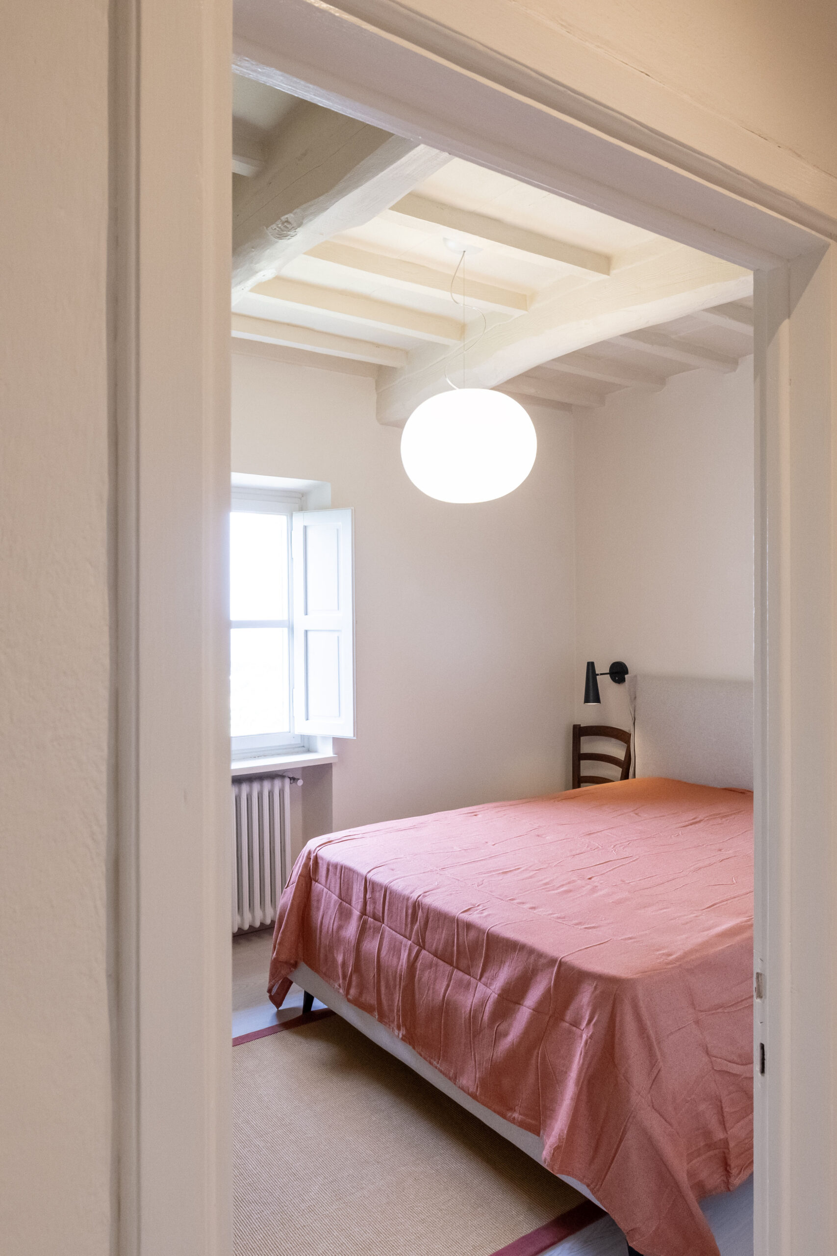 Room view of a pink-bedroom with a hanging spherical ceiling light, exposed white ceiling beams, and an open window letting in daylight.