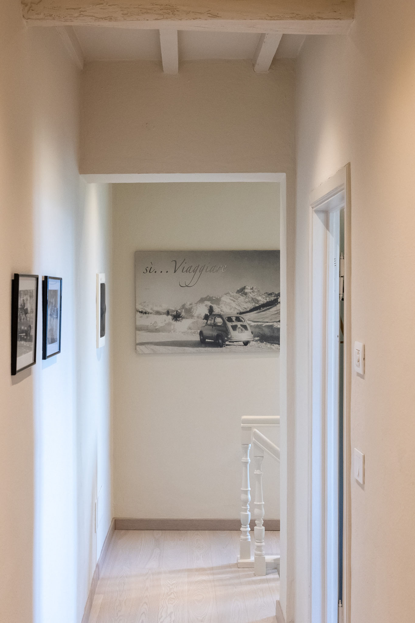 Narrow bright hallway with light wooden floor, white walls and exposed ceiling beams; a large black-and-white landscape print with a vintage car hangs on the far wall.