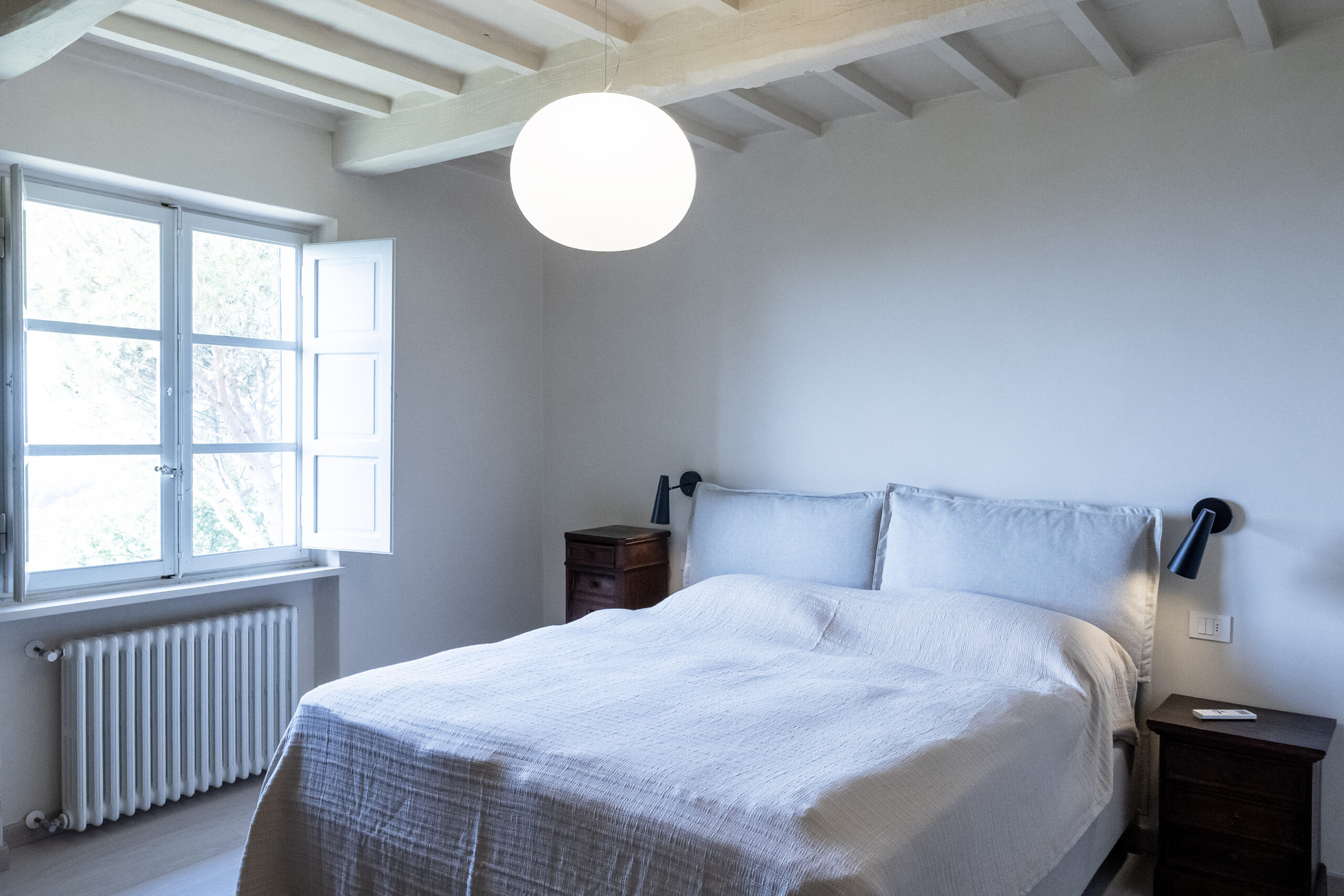 Bright bedroom with pale blue walls, exposed white beams, a large round pendant light, and an open window beside a neatly made bed with wooden nightstands nearby.
