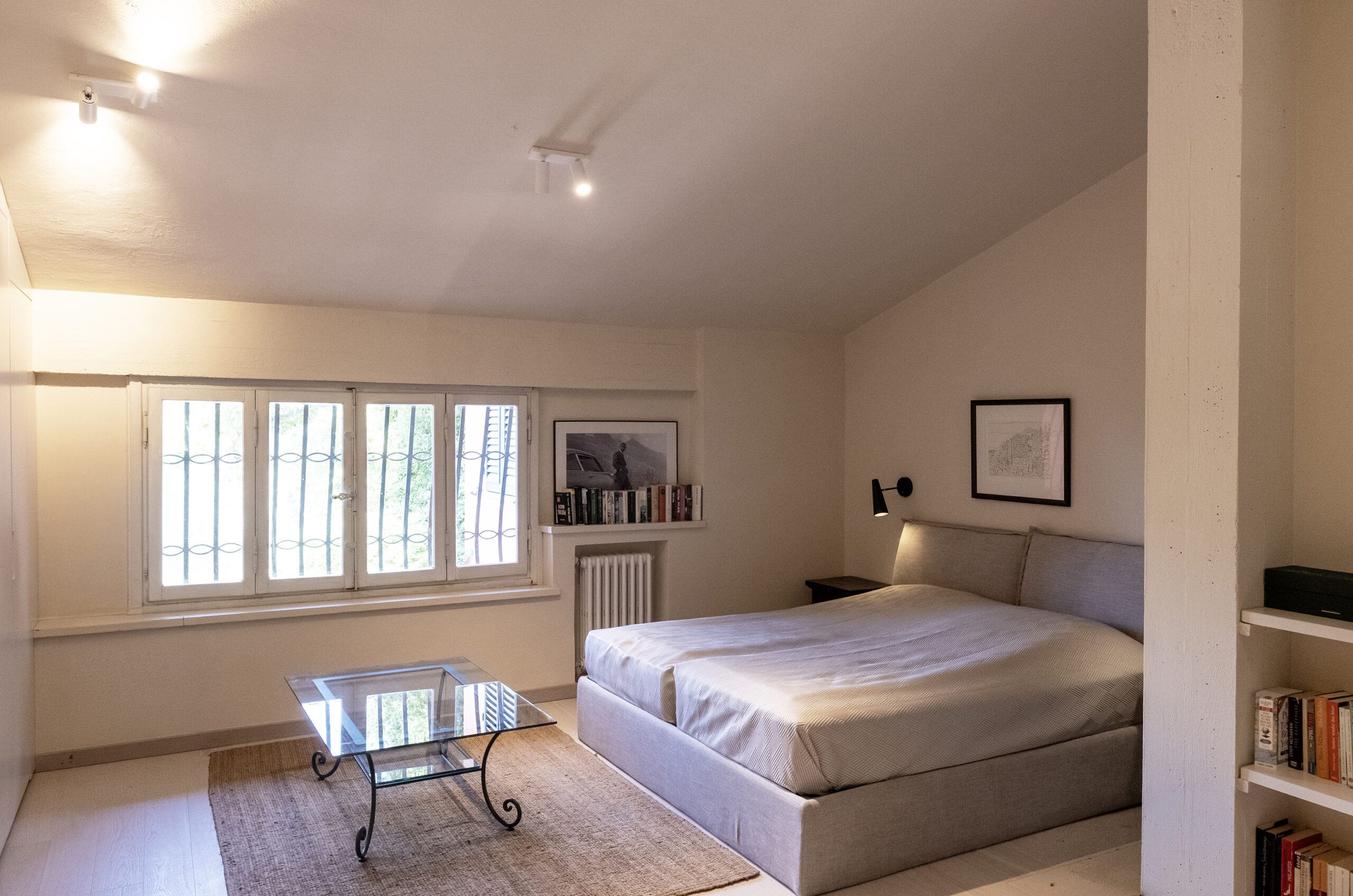 Cozy beige bedroom with a large bed, glass coffee table on a rug, and a window with bars along the wall.