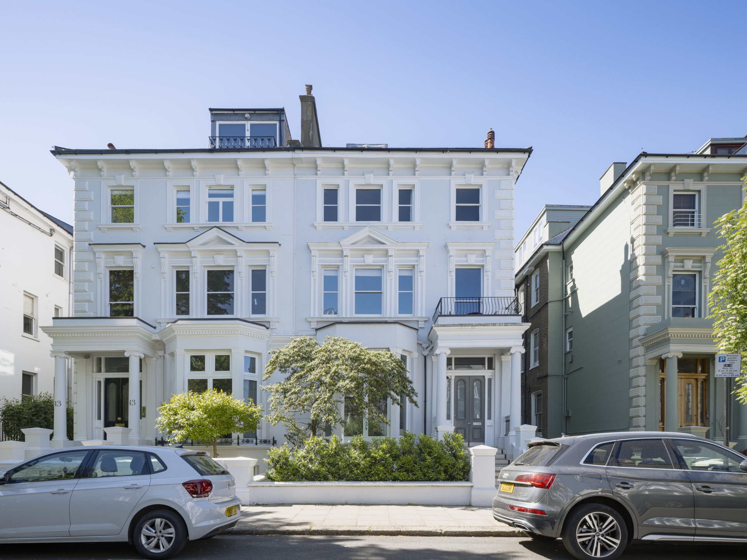 White, ornate townhouses on a sunny street with trimmed hedges and parked cars in front.