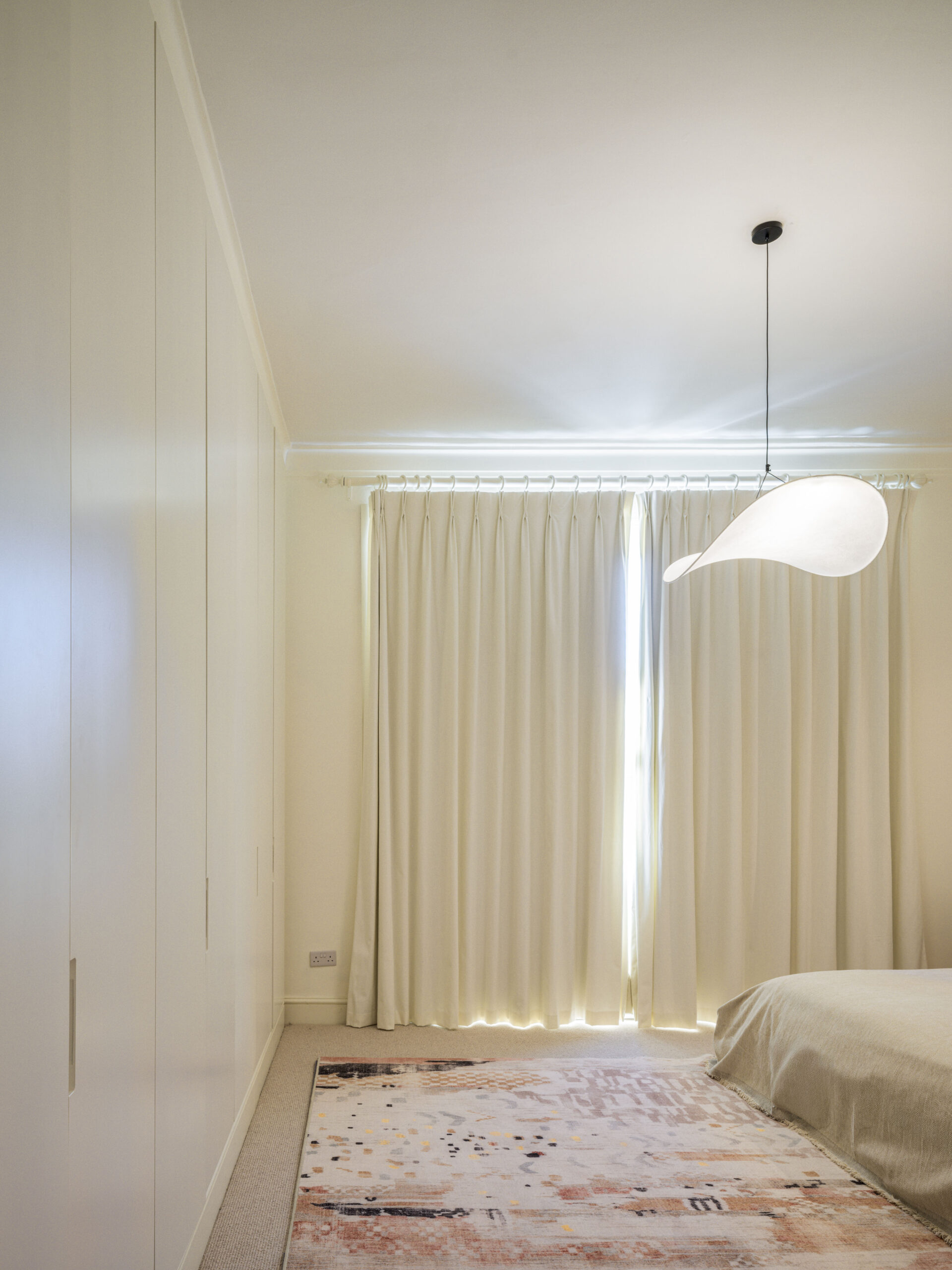 Bright minimalist bedroom corner with a curved pendant light, white walls, cream curtains, and a pale patterned rug left of a bed edge.