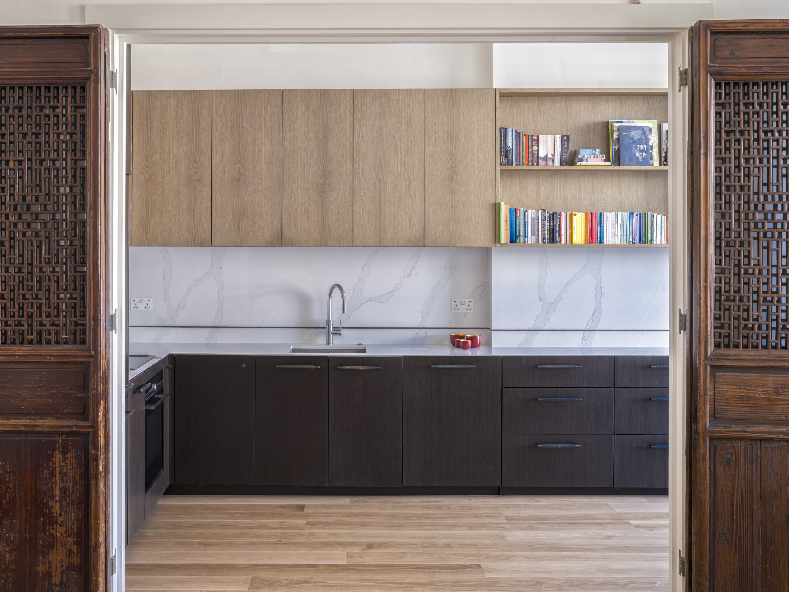 Kitchen seen through carved wooden doors: light wood upper cabinets, dark lower cabinets, marble backsplash, and a bookshelf on the right.
