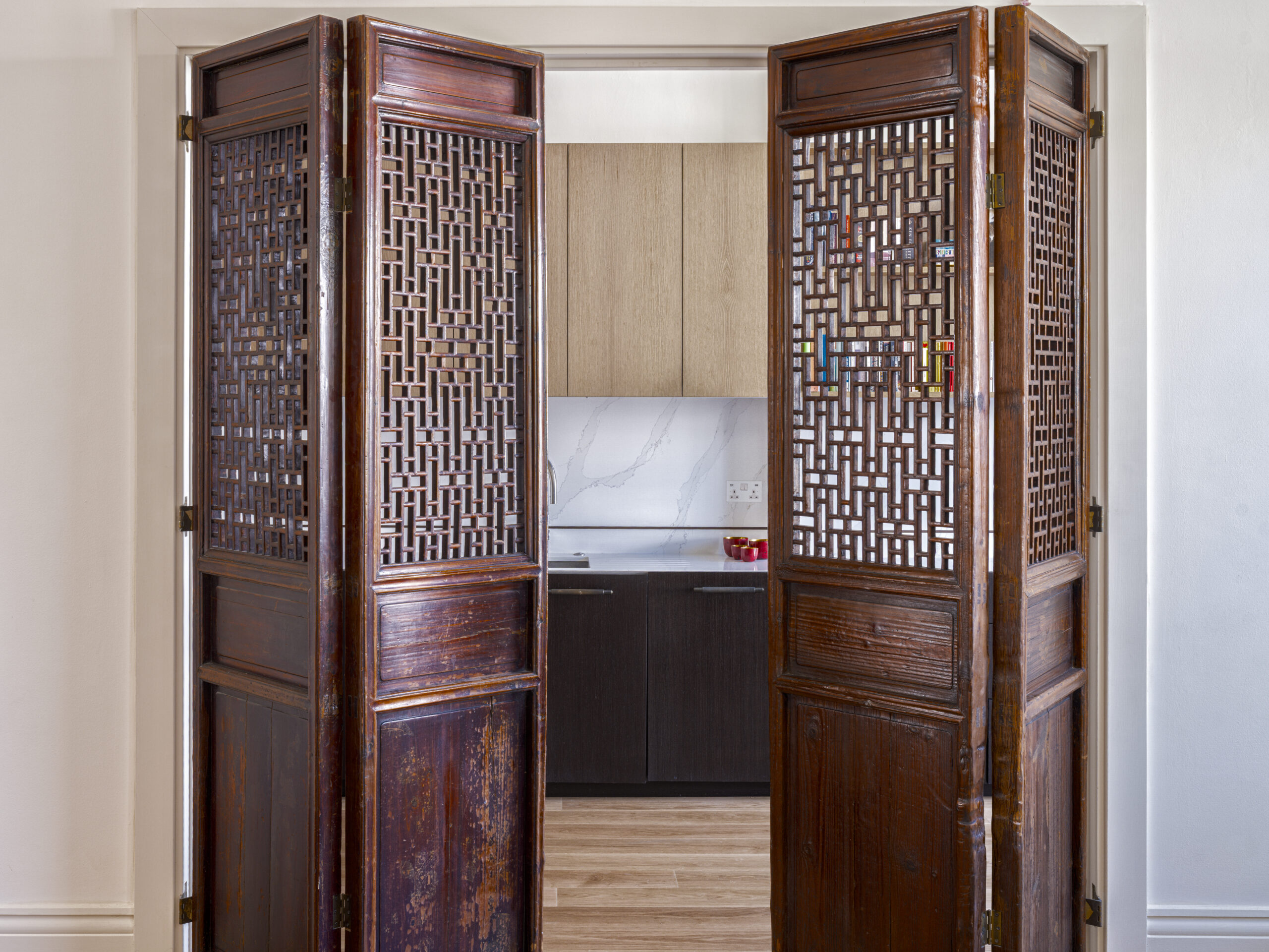 Open carved wooden folding doors revealing a modern kitchen with beige cabinets and marble backsplash inside.