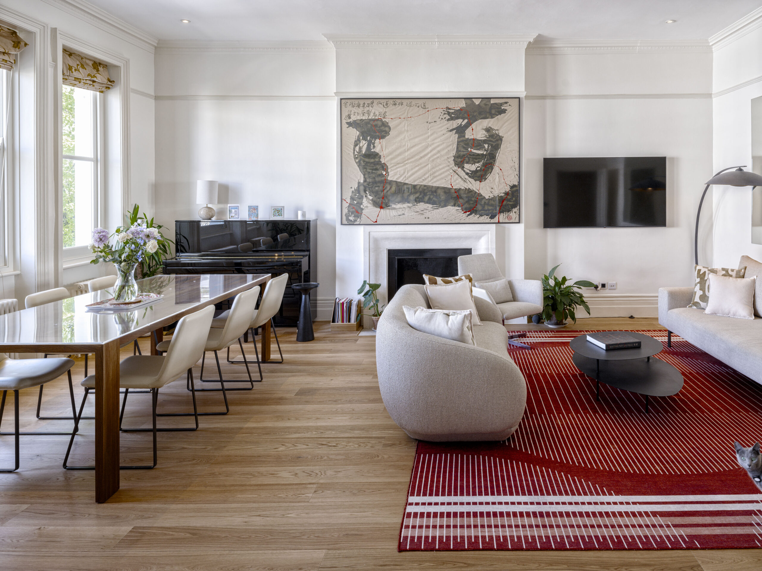 Bright living room with a red striped rug, white walls, and a glass dining table with cream chairs; a piano against the left wall and a vase of flowers on the table.