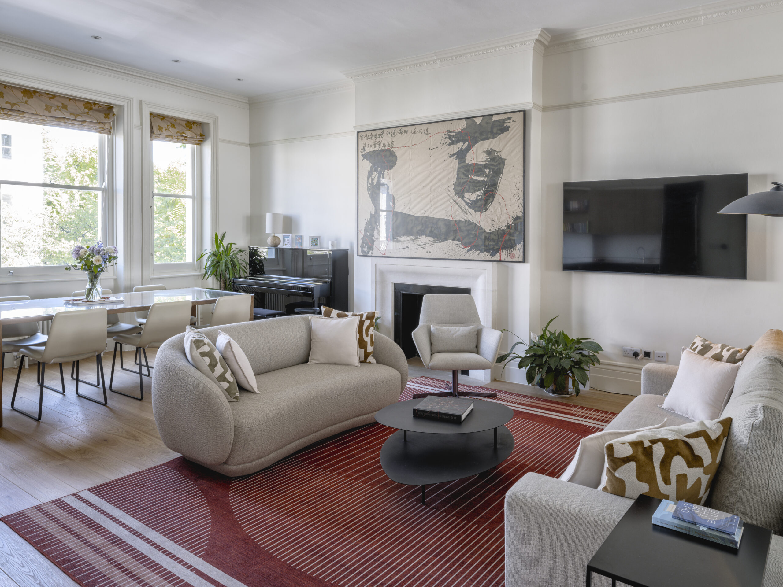 Bright living and dining room with beige sofas, a black round coffee table, and a red striped rug; large artwork above the fireplace and a wall-mounted TV to the right.