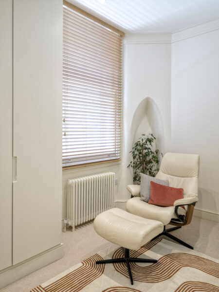Cozy reading nook with beige lounge chair, matching ottoman, and a potted plant by a window with wooden blinds.