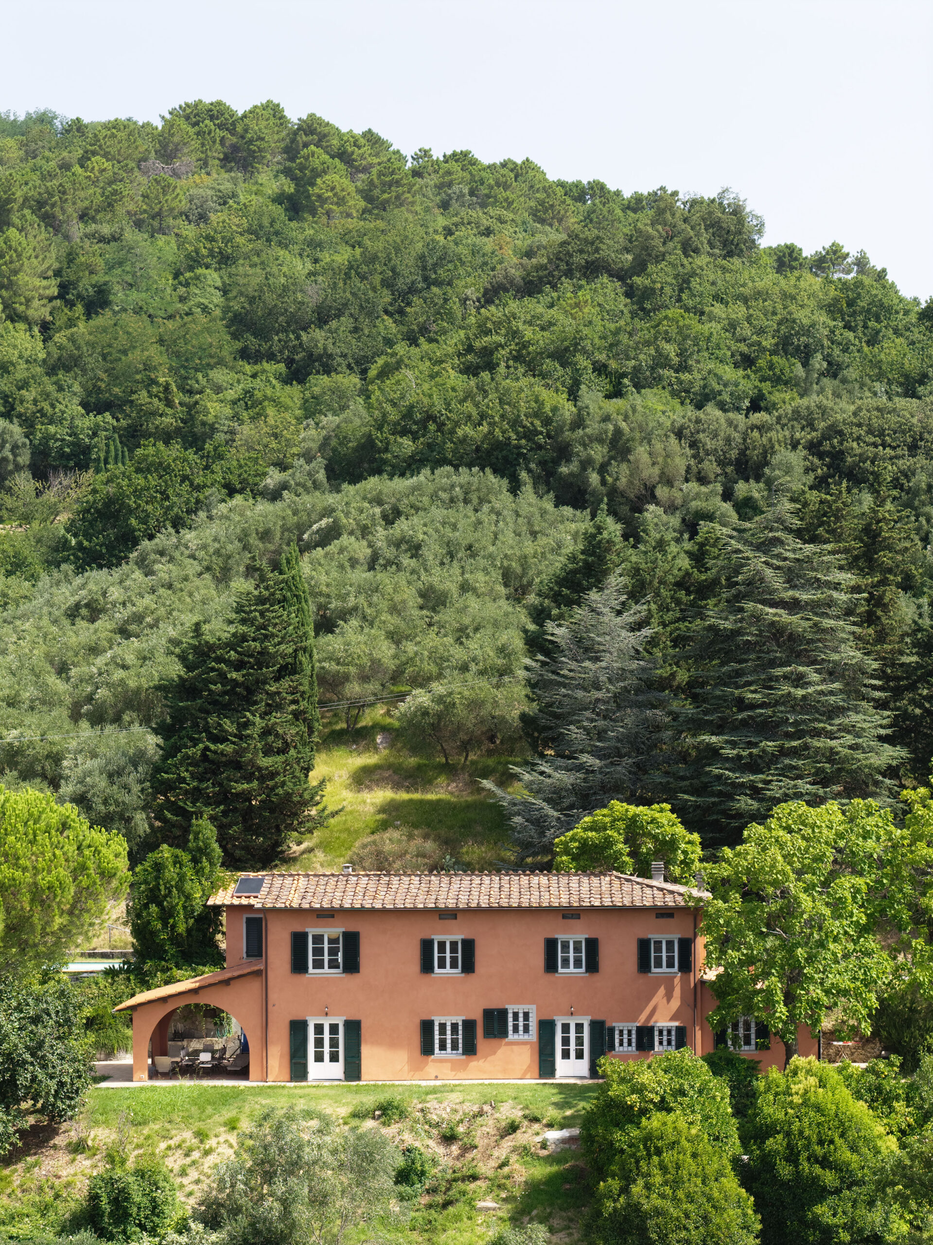Orange villa with green shutters at the edge of a grassy yard, backed by a dense forested hillside of pines and deciduous trees