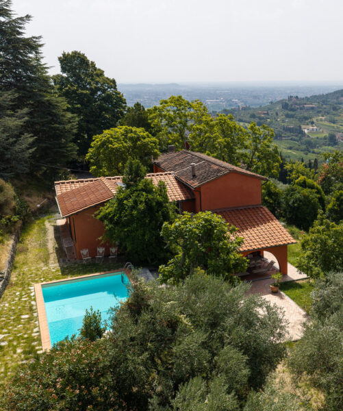 Terracotta villa with tiled roof beside a turquoise rectangular pool on a hillside among trees, overlooking a valley.