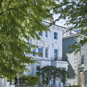 White townhouse façade with decorative trim visible through green tree branches under a blue sky.