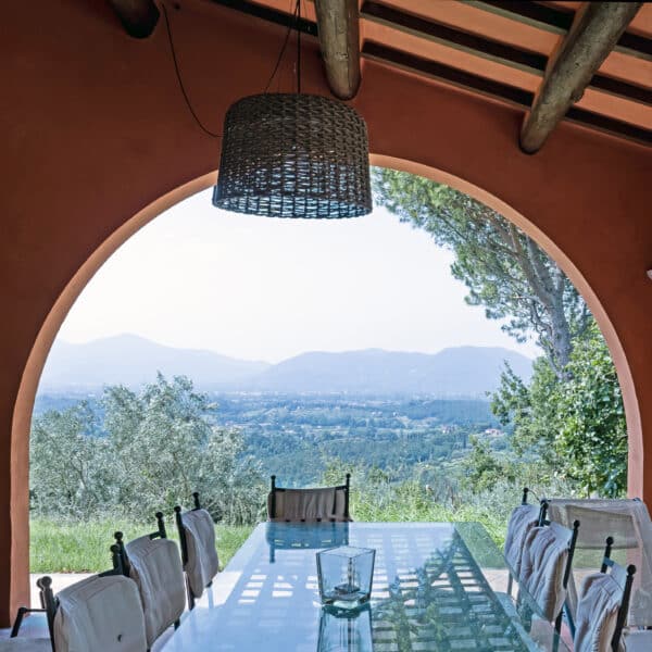 Outdoor dining area with a glass table and cushioned chairs under a terracotta veranda, overlooking mountains and trees in the distance.",