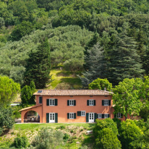 Terracotta two-story villa with green shutters, tiled roof, and a shaded archway, set in a lush, tree-filled landscape.